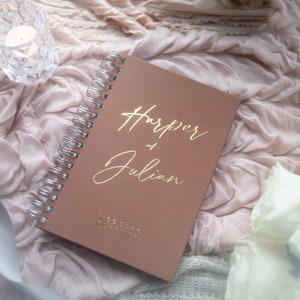 May include: A blush pink wedding planner with a gold spiral binding. The cover has the names "Harper & Julian" written in gold script and the words "Wedding Planner" in gold lettering.