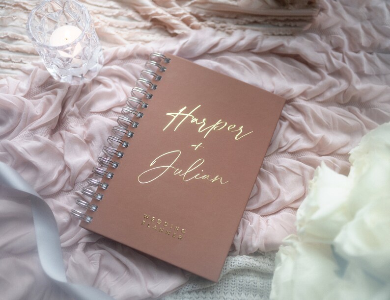May include: A blush pink wedding planner with a gold spiral binding. The cover has the names "Harper & Julian" written in gold script and the words "Wedding Planner" in gold lettering.