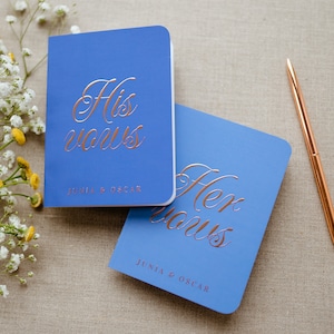 May include: Two blue notebooks with gold foil lettering. The top notebook says "His vows" and the bottom notebook says "Her vows". Both notebooks have the names "Junia & Oscar" printed on them.
