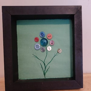 May include: A framed artwork featuring a flower design made from colorful buttons. The flower has a blue glass marble center, with a green background and a dark brown frame. The stem and branches are painted green.