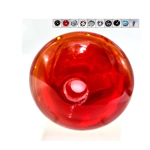 May include: A red glass bead with a small hole in the center. The bead has a slight orange hue and is translucent.