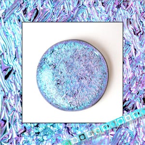 May include: A round, dichroic glass cabochon with a textured surface. The cabochon features a blend of blue, purple, and iridescent colors. The background is a textured, iridescent foil in shades of blue, purple, and silver.