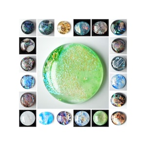 May include: A collection of dichroic glass cabochons in various colors and designs. The assortment includes round, iridescent pieces with shades of blue, green, gold, and purple. The central piece is a large, round, light green cabochon with gold glitter.