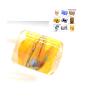 May include: A yellow glass bead with blue and brown swirls. The bead is rectangular in shape and has a glossy finish.