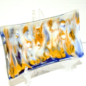 Puede incluir: Un plato rectangular de vidrio con un diseño marmoleado azul, blanco y dorado. El plato está sobre un soporte de plástico transparente.
