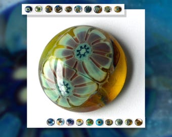 May include: A round glass bead with a floral design. The bead is yellow, brown, and blue with white flowers.