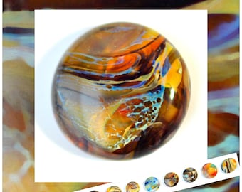 May include: A round, glass paperweight with a swirling design of orange, yellow, blue, and brown colors. The paperweight has a glossy finish and is set against a white background. A selection of similar paperweights are shown below.