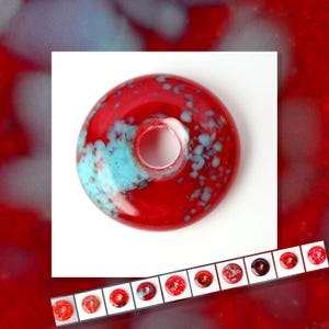 Puede incluir: Una cuenta de vidrio roja con un diseño moteado turquesa y blanco. La cuenta tiene forma de rosquilla con un agujero central. Se muestra una fila de cuentas similares debajo.