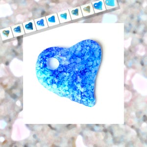 May include: A heart-shaped, blue and white resin pendant with a hole. The pendant has a textured, speckled appearance. Above, a row of small heart-shaped pendants in various shades of blue and gold are displayed on a white background.