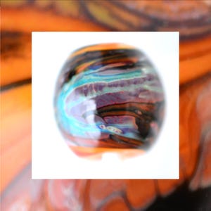 May include: A colorful, round glass bead with a swirling pattern of teal, purple, orange, and black. The bead has a glossy finish and is set against a white background. The bead is a decorative element.