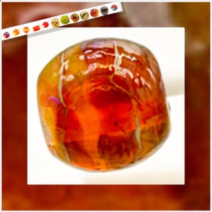 May include: A close-up of a large, irregularly shaped, amber-colored glass bead. The bead has a glossy finish with internal streaks of red and yellow. The background is white, with a brown border.