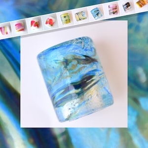 May include: A cylindrical glass object with a blue and green marbled design. The glass has a translucent quality, with darker streaks and bubbles visible within the material. Several smaller, colorful glass beads are arranged above the main object.