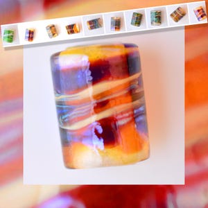 Puede incluir: Una gran cuenta de vidrio cilíndrica con un patrón arremolinado de tonos naranja, amarillo y púrpura. La cuenta tiene un acabado brillante y está rodeada de cuentas más pequeñas de varios colores. El diseño recuerda al vidrio fundido.