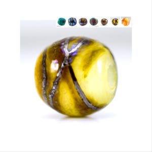 Puede incluir: Una cuenta de vidrio amarilla con rayas negras y plateadas. La cuenta tiene un acabado brillante y es redonda.