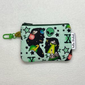 May include: A small, mint green zippered pouch with an alien-themed pattern. The design features silhouettes of figures with space-themed elements, stars, and the letters "X" and "Y". A green clip and a tag that says "hello Orchid" are attached.