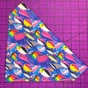 May include: Triangular blue bandana with a repeating pattern of colorful fish. The fish feature rainbow, pink, yellow, black, and white designs. The bandana is likely made of fabric and is suitable for pets.