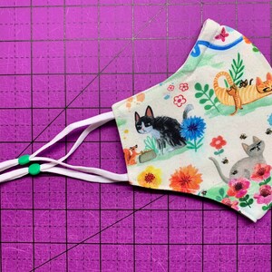 May include: A white face mask with a colorful pattern of cats, flowers, and bees. The mask has white elastic straps with green toggles.