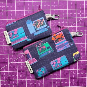 May include: Two small zippered pouches with a dark blue background and a repeating pattern of retro computers with flowers in the screen. The pouches have pink zippers and a pink fabric tag with the text "hello orchid" on them.