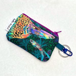 Op de afbeelding: Een kleine etui met ritssluiting met een levendig visontwerp. De etui heeft een donkergroene achtergrond met kleurrijke visillustraties. De rits is paars en een klein label leest "hello orchid". Een blauwe riem met een sleutelhanger is bevestigd.
