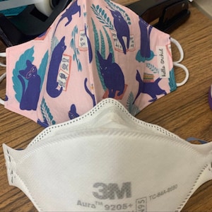 May include: Two face masks. One is a pink fabric mask with a pattern of blue cats, fish, and plants. The other is a white 3M Aura 9205+ respirator mask. The fabric mask has the text "hello orchid".