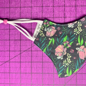 May include: A black face mask with a floral print featuring pink flowers, green leaves, and white dots. The mask has white ties and a pink adjustable nose wire. The text "Hello Orchid" is printed on the mask.