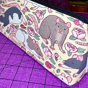 May include: A yellow fabric pencil case with a repeating pattern of rats and pink flowers. The case has a black zipper and a white tag with the text "Little Orchid".