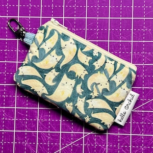 Puede incluir: Una pequeña bolsa con cremallera de fondo crema y estampado de gatos azules. La bolsa tiene una cremallera azul claro y un llavero plateado. La bolsa tiene una etiqueta blanca con el texto "hello orchid" impreso en ella.