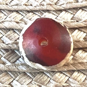 May include: A round, red bead with a hole in the center, set against a woven, beige background. The bead has a white, irregular edge. The bead's color transitions from a deep red to a lighter shade.