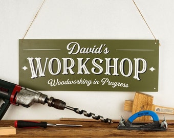 May include: Green metal sign with white text that reads "David's Workshop - Woodworking in Progress". The sign is hanging from a string and there are various woodworking tools in front of it, including a drill, a hammer, and a sander.