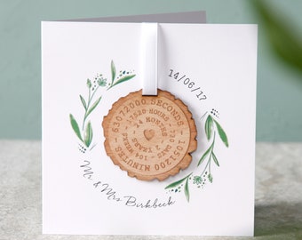 May include: A white card with a green floral design and a wooden circle attached with a white ribbon. The circle has the text "1051200 MINUTES - 104 WEEKS - 2 YEARS - 730 DAYS - 17520 HOURS - 63072000 SECONDS" and a small heart. The card also has the text "14/06/17" and "Mr & Mrs Birkbeck".