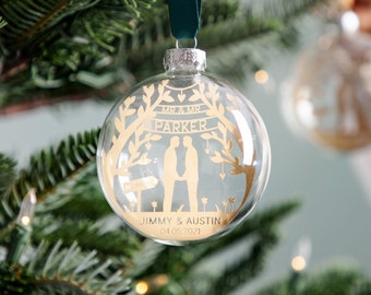 May include: A clear glass Christmas bauble with a gold design of two figures holding hands in front of a tree. The bauble also has the text "MR & MR PARKER" and "JIMMY & AUSTIN 04.05.2021".