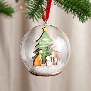 May include: A clear glass Christmas ornament with a wooden cutout of a Christmas tree, a fox, a rabbit, and a deer. The tree has the text "Finley Alistair Smith 22.09.2023 First Christmas" on it.