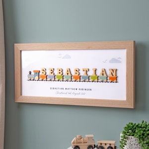 May include: A framed print with a wooden train carrying the name "SEBASTIAN" in colorful letters. The train is on a white background with clouds and birds. The text "SEBASTIAN MATTHEW ROBINSON Christened 3rd August 2022" is below the train.