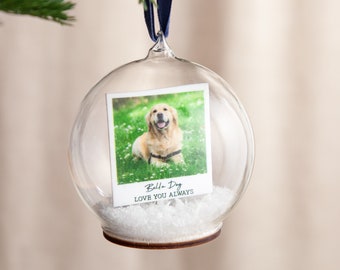 May include: A clear glass Christmas ornament with a photo of a golden retriever inside. The photo is a polaroid style with the text "Bella Dog LOVE YOU ALWAYS". The ornament is hanging from a navy blue ribbon.