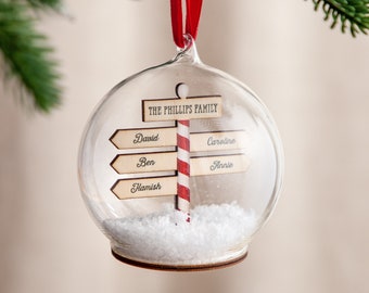 May include: A clear glass Christmas ornament with a snow-filled base. Inside the ornament is a wooden signpost with the text "THE PHILLIPS FAMILY" at the top and the names "David", "Ben", "Hamish", "Caroline", and "Annie" on the signposts.