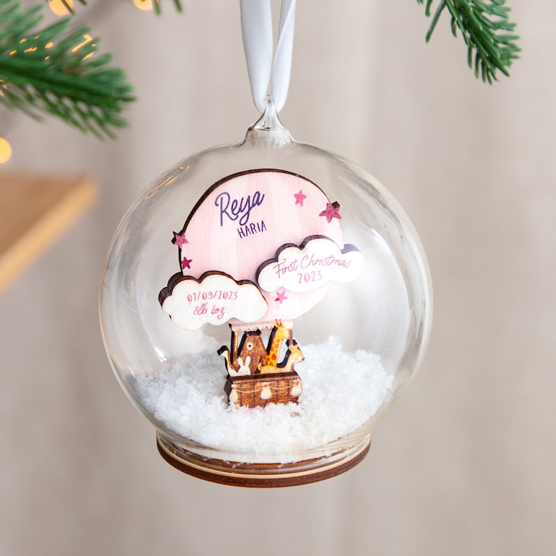 May include: A glass ornament with a pink and white wooden hot air balloon inside. The balloon has the name "Reya Maria" and the date "01/03/2023" written on it. The ornament also has the text "First Christmas 2023" on it.