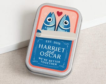 May include: A silver rectangular tin with a sardine can design. The lid features two blue fish and a red heart. The tin reads "Harriet and Oscar" and "We're Better Together."