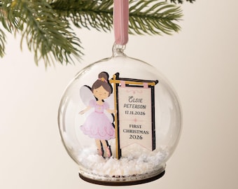 May include: A clear glass spherical ornament containing a fairy illustration and a sign with the text Elsie Peterson 17.11.2026 First Christmas 2026. The base is filled with white faux snow and the ornament hangs from a light pink ribbon.