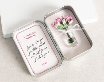 May include: An open silver tin with a miniature bouquet of pink flowers in a small glass vase, secured with a white ribbon. The left side of the tin features a card with handwritten text: "A NOTE from DELILAH. Like my love for you, these flowers will last forever. I adore you x. 2026."