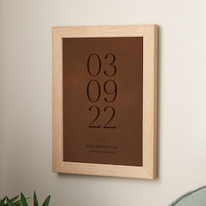 May include: A framed print with a brown background and the date 03 09 22 in large, dark text. Below the date is the text "MR & MRS DOUGAL Leather Anniversary" in a smaller font. The frame is made of light-colored wood.