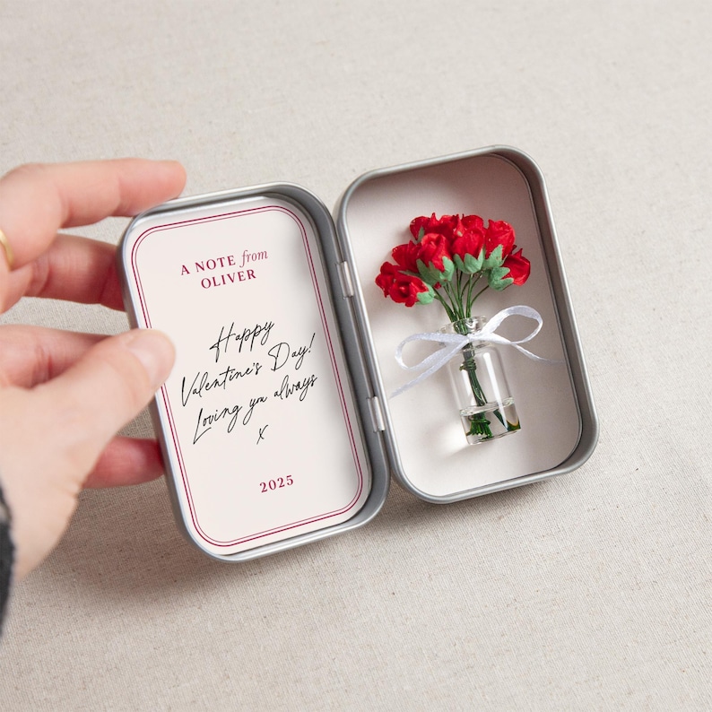 May include: A small, open silver tin containing a miniature bouquet of red roses in a tiny glass vase tied with a white ribbon. The left side of the tin has a card with the text "A NOTE from OLIVER" and "Happy Valentine's Day! Loving you always X 2025."