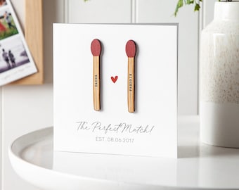 May include: A white card with two wooden matchsticks with red tops. The matchsticks have the names "SASSIA" and "FREDDIE" written on them. A small red heart is between the matchsticks. The card reads "The Perfect Match! EST. 08.06.2017".