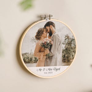 May include: A round, wooden embroidery hoop holding a printed photo of a couple embracing. The photo is of a wedding, with the text "Mr & Mrs Byrd Est. 2023" printed below the image.