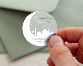 May include: A silver sticker featuring a crescent moon and mosque silhouette design. The text "Ramadan Mubarak" is written in a cursive font. The sticker also includes the text "THE HUSSAIN FAMILY".