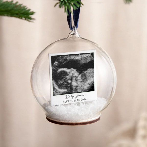 May include: A clear glass bauble containing a black and white ultrasound image. The bauble has a white base with the text "Baby Jones CHRISTMAS 2024" printed on it.