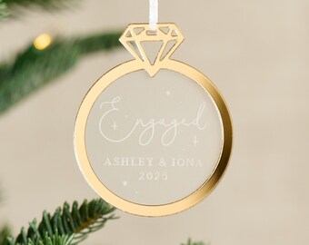 May include: A gold-coloured engagement ring Christmas tree ornament. The word "Engaged" is written in cursive, with the names "Ashley & Iona" and the year "2025" below. It hangs from a white ribbon.