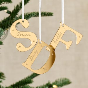 May include: Gold-coloured Christmas tree decorations in the shape of the letters S, D, and F, each with a name engraved: Spencer, Daisy, and Freddie. They hang from white ribbons and are decorated with small star shapes.