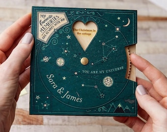 May include: A teal blue wooden card with a heart-shaped window revealing the text "The Christmas in the cottage". The card has a spinning wheel with the text "My favourite MEMORIES ARE ALWAYS WITH YOU" and "YOU ARE MY UNIVERSE". The card is personalized with the names "Sara & James".