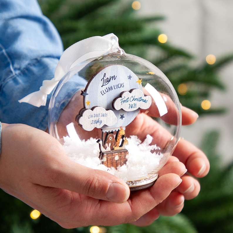 May include: A clear glass Christmas ornament with a wooden hot air balloon inside. The balloon has a blue and white cloud with the text "First Christmas 2021" and a smaller cloud with the text "17th July 2021 2.8kg". The balloon is carrying a giraffe, a bear, and a penguin. The ornament also has a white ribbon and the text "Liam Elijah Lucas" on a blue and white cloud.