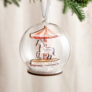 May include: A clear glass Christmas ornament with a wooden carousel inside. The carousel has a red and white striped canopy and a white horse with a blue mane and tail. The ornament also includes the text "First Christmas 2023", "Madeleine Mildred", "14.08.2023", and "6lb 10oz".